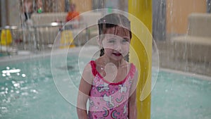 A little girl with wet hair in a pink swimsuit stands in the rain in the water park