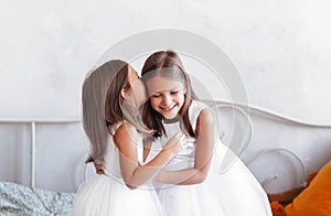 Little girl whispers in her sister`s ear. Two girls girlfriends in a light room