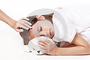 A little girl in a white dress sleeps with her favorite toy on a white background, and mom `s hand irons her on the head.