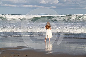 Little Girl in White Dress Standing at Ocean Edge as Wave Rolls In