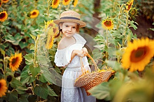 Little girl in a white dress, a straw hat with a basket full of sunflowers smiling at the camera in a field of