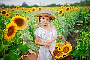 Little girl in a white dress, a straw hat with a basket full of sunflowers smiling at the camera in a field of