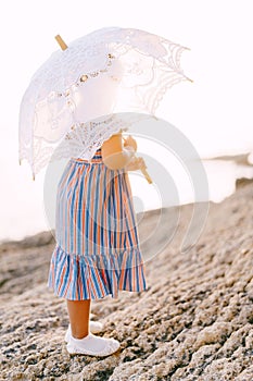 Little girl with a white openwork sun umbrella stands on the seashore