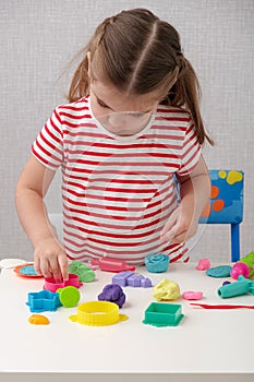 Little girl in white and red striped T-shirt is enthusiastically playing with plasticine, play dough, playdough, mass for modeling