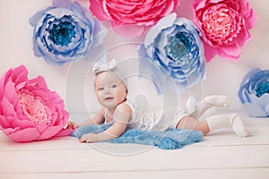 Little girl in a white suit and white socks in a white room with bright pink and blue paper flowers, baby photo session