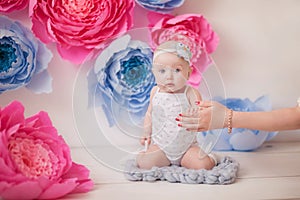 Little girl in a white suit and white socks in a white room with bright pink and blue paper flowers, baby photo session