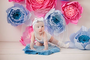 Little girl in a white suit and white socks in a white room with bright pink and blue paper flowers, baby photo session