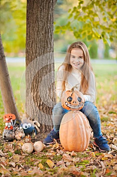 Little girl in a white sweater and jeans on a background of green textural natural background. The girl sits on pumpkins and laugh