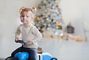 Little girl in a white sweater rides a toy vintage blue car. Christmas tree in the background, gifts, lights