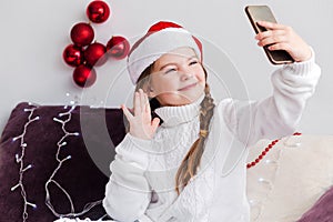 A little girl in a white sweater and Santa Claus hat looks at a mobile phone and says hello. Girl talking by video call