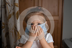Little girl  in a white T-shirt covers her face with her hands in a medical mask at home