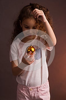 A little girl in a white t-shirt with curly long hair, holding a ball of Christmas tree decoration, looks away on a dark purple