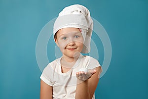 Little girl with a white towel on her head washes her face on a blue background. The child has foam on his hand.