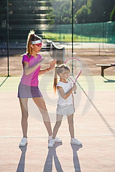 Little girl in white uniform hitting ball under net during tennis training with coach. Attractive female coach making