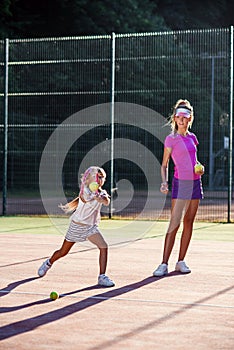 Little girl in white uniform hitting ball under net during tennis training with coach. Attractive female coach making