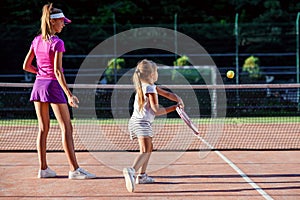 Little girl in white uniform hitting ball under net during tennis training with coach. Attractive female coach making