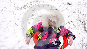 Little girl in winter in bright warm clothes rides on a plastic blue sled