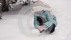 Little girl in winter park sculpts snowman. From the sky falling snow.