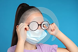 Little girl wiping foggy glasses caused by wearing medical face mask on blue background. Protective measure during coronavirus