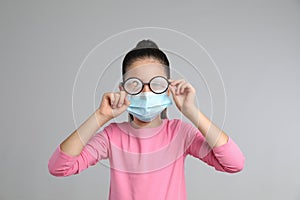 Little girl wiping foggy glasses caused by wearing medical face mask on grey background. Protective measure during coronavirus