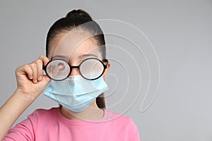 Little girl wiping foggy glasses caused by wearing medical face mask on grey background, space for text. Protective measure during