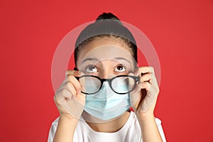 Little girl wiping foggy glasses caused by wearing medical face mask on red background. Protective measure during coronavirus