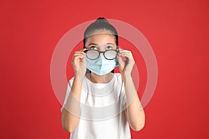 Little girl wiping foggy glasses caused by wearing medical face mask on red background. Protective measure during coronavirus