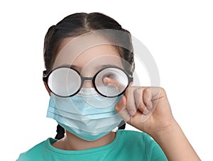 Little girl wiping foggy glasses caused by wearing medical face mask on white background. Protective measure during coronavirus