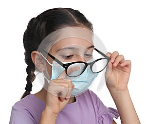 Little girl wiping foggy glasses caused by wearing medical face mask on white background. Protective measure during coronavirus