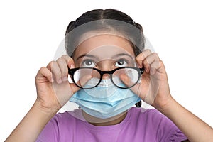 Little girl wiping foggy glasses caused by wearing medical face mask on white background. Protective measure during coronavirus