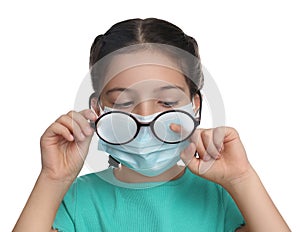 Little girl wiping foggy glasses caused by wearing medical face mask on white background. Protective measure during coronavirus