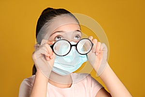 Little girl wiping foggy glasses caused by wearing medical face mask on yellow background. Protective measure during coronavirus