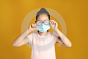 Little girl wiping foggy glasses caused by wearing medical face mask on yellow background. Protective measure during coronavirus