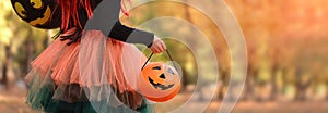 Little girl in witch costume with a balloon and a pumpkin bucket playing in autumn park. Child having fun at Halloween trick or