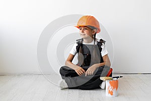 Little girl in work clothes of a builder and an orange hard hat sits on the floor. Parents help concept.