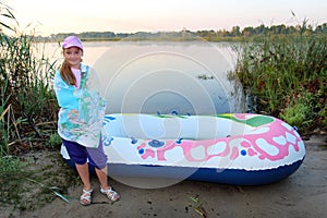 little girl wrapped in a towel stands on the river bank near an inflatable boat