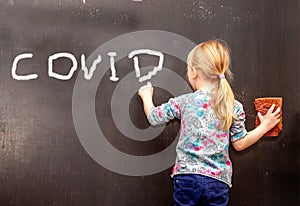Little girl writing COVID on black chalkboard