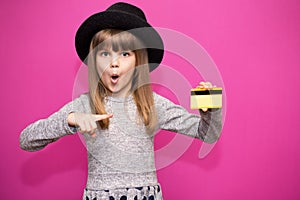 Little girl 6-7 years old in black hat with shoked face hold credit bank card isolated on pink. Childhood lifestyle concept photo