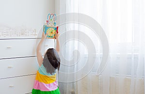 A little girl 3 years stretches to get an educational toy from a high shelf.