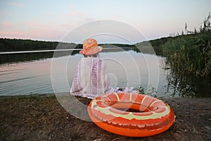 A little girl in a yellow hat, covered with a towel, sits on the shore of the lake, next to an inflatable circle, summer evening-