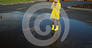 little girl in yellow raincoat and yellow rubber boots is using smartphone