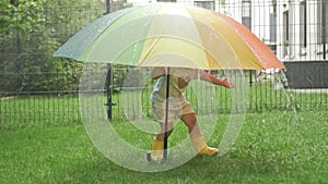 Little girl in yellow rubber boots under a large colorful umbrella in the midst of summer rain.