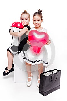 2 little girls with elegant dresses sit with large bags with heart-shaped balloons inside