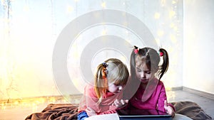 Little girls involved in use of tablet and sit on floor in bright room with garland on wall.