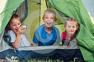 Little girls sisters and brother boy sitting inside green camp tent drinking tea and cheerfully laughing and looking into camera.