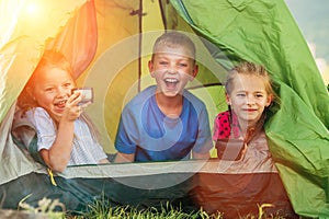 Little girls sisters and brother boy sitting inside green camp tent drinking tea and cheerfully laughing and looking into camera.