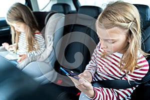 Little girls, sisters are driving in car, children play video games in tablet and mobile phone. Traveling on road in safe baby