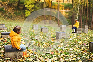 Little girls sisters in yellow jacket and raincoat sit on concrete posts of the foundation in the autumn forest or park. Maple