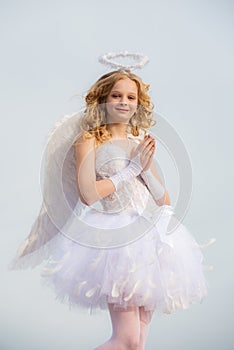 Little goddess with white wings alone on blue sky background. Portrait of a cupid little girl pray. A beautiful teen
