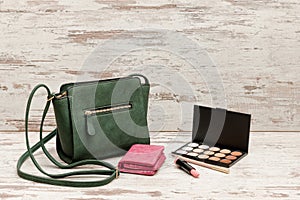 Little green ladies handbag, pink purse, eyeshadow palette and lipstick on wooden background. fashion concept